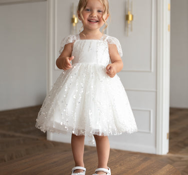Little girl wearing a luxury white sequin flower girl dress by Aylu Bebe, featuring flutter sleeves and a sparkling tulle skirt, standing in bright modern room. Perfect for weddings, parties and special occasions.