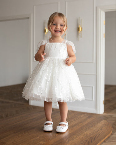 Little girl wearing a luxury white sequin flower girl dress by Aylu Bebe, featuring flutter sleeves and a sparkling tulle skirt, standing in bright modern room. Perfect for weddings, parties and special occasions.