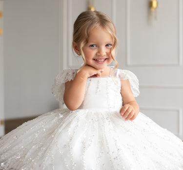 Little girl wearing the Dahlia Blossom white sequin flower girl dress by Aylu Bebe, sitting on wooden floor and smiling