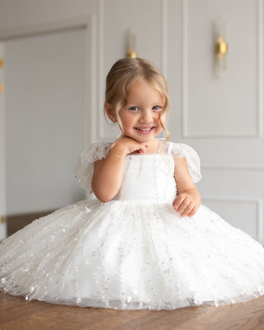 Little girl wearing the Dahlia Blossom white sequin flower girl dress by Aylu Bebe, sitting on wooden floor and smiling