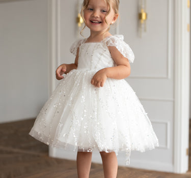 Little girl wearing a luxury white sequin flower girl dress by Aylu Bebe, featuring flutter sleeves and a sparkling tulle skirt, standing in bright modern room. Perfect for weddings, parties and special occasions.