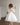 Little girl wearing a luxury white sequin flower girl dress by Aylu Bebe, featuring flutter sleeves and a sparkling tulle skirt, standing in bright modern room. Perfect for weddings, parties and special occasions.