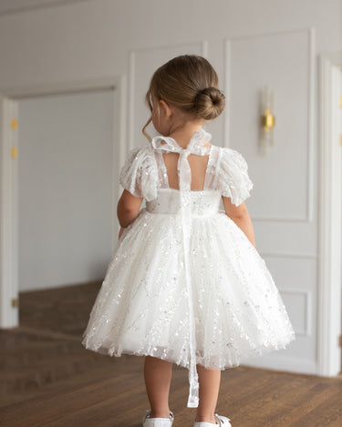 Little girl wearing a luxury white sequin flower girl dress by Aylu Bebe, featuring flutter sleeves and a sparkling tulle skirt, standing in bright modern room. Perfect for weddings, parties and special occasions.