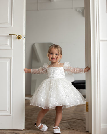 Full body front view of child wearing luna sequin dress, standing against white wall