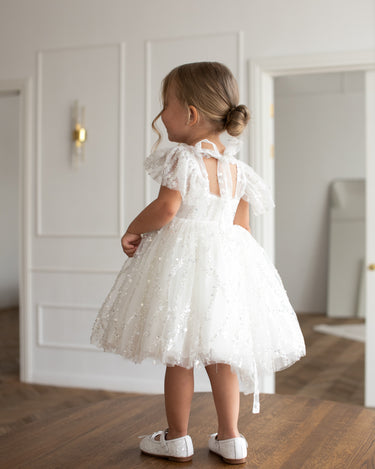 Young girl wearing the Dahlia blossom white sequin flower girl dress with flutter sleeves, standing near a doorway on a wooden floor.