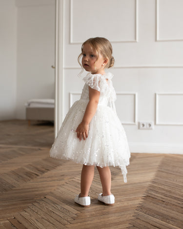 Little girl wearing the Dahlia Blossom white sequin dress by Aylu Bebe, standing on a wooden floor and looking back