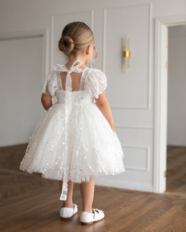 Back view of a young girl wearing the Dahlia Blossom white sequin flower girl dress with flutter sleeves and a bow tie detail, standing on a wooden floor.