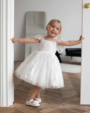 Little girl wearing the Dahlia Blossom white sequin flower girl dress by Aylu Bebe, smiling while holding a door frame and twirling. 